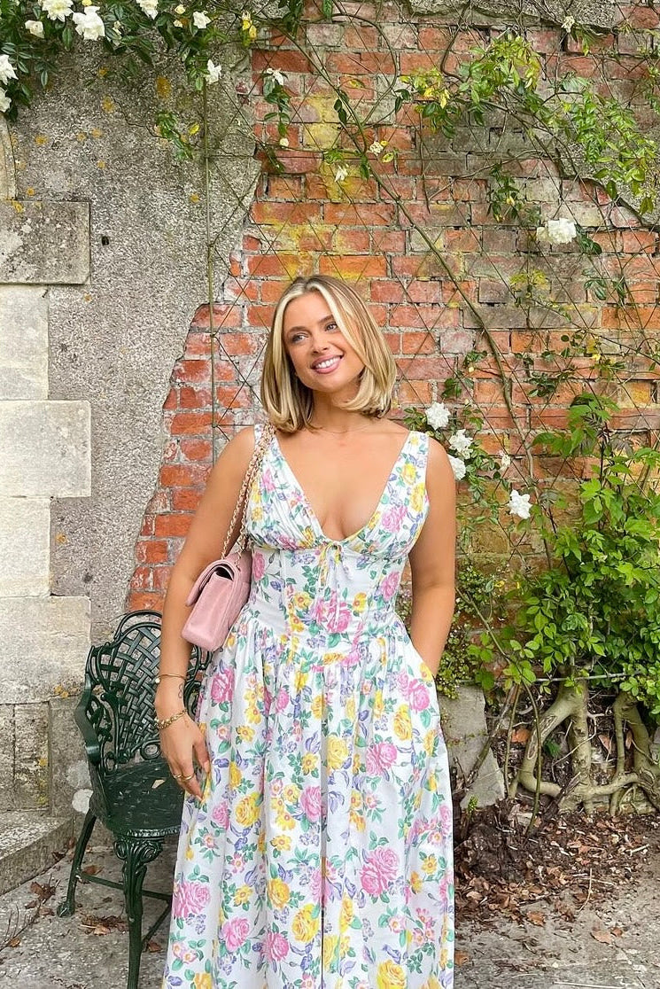 Smiling woman in a white floral midi dress with a pink shoulder bag, standing outdoors against a brick and vine wall.
