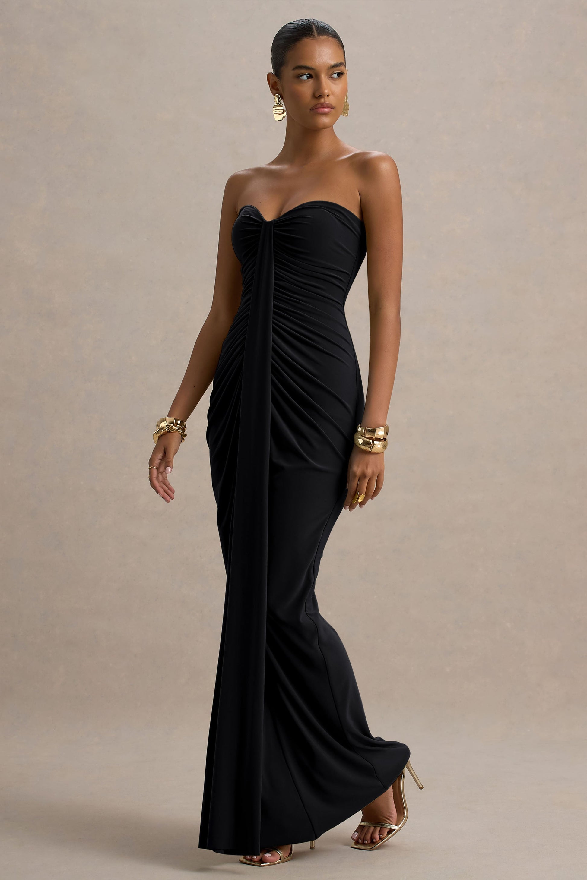 Side view of a black strapless ruched maxi dress with a long flowing skirt and gold heels.