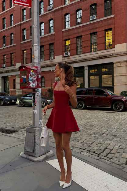 Red Lucy skater mini dress with thick straps and a structured skirt, worn with white pointed heels and a white quilted bag.