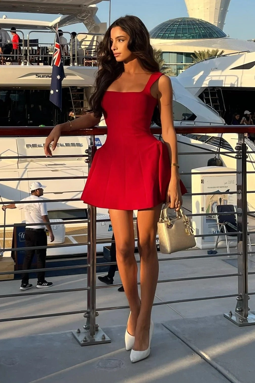 Red Lucy skater mini dress with a square neck and a flared skirt, paired with white heels and a gold top-handle bag on a yacht.