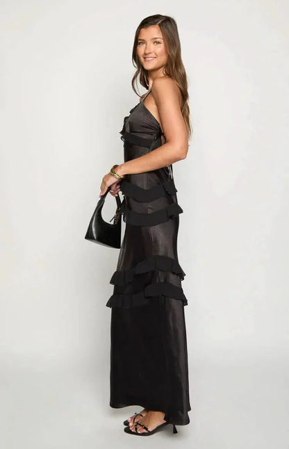 Side view of a woman wearing a black Madeline ruffle maxi dress with spaghetti straps, holding a black handbag, showcasing the tiered ruffles.