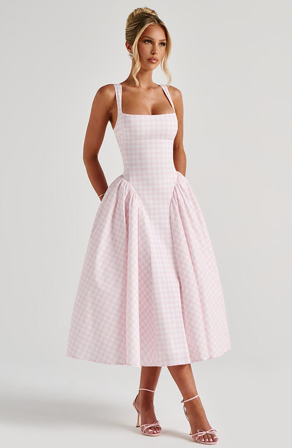 Pink gingham midi dress with wide straps and a cinched waist, flowing into a full skirt, shown with pink heels.