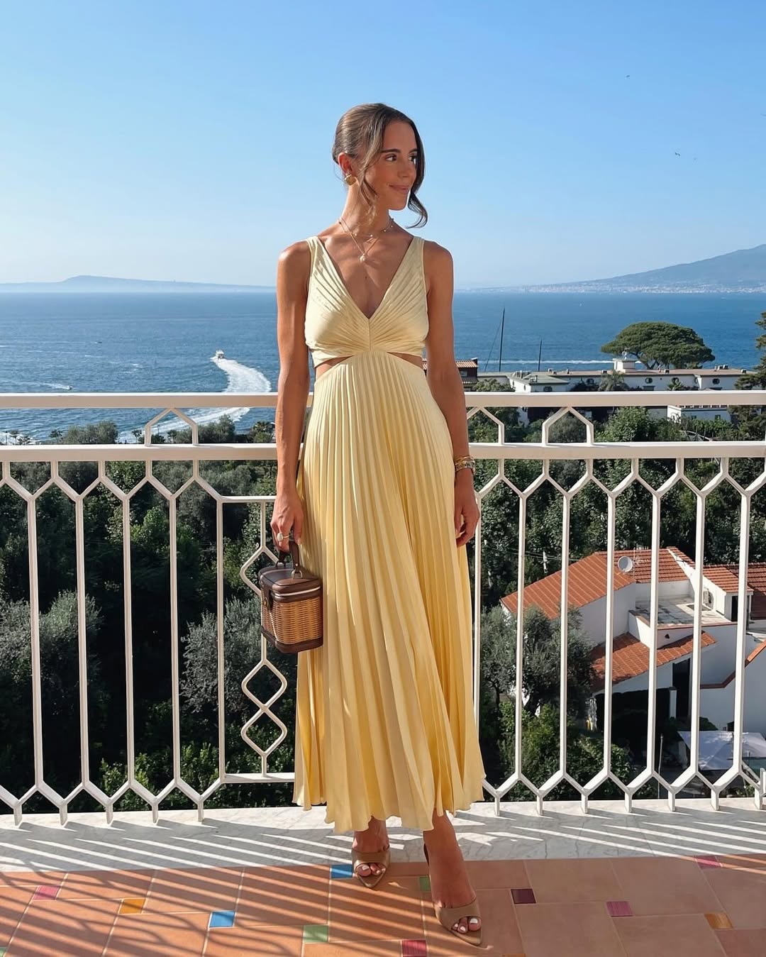 Maria Pleated Maxi Dress in light yellow with a deep V-neckline and side cut-outs, standing on a balcony overlooking the sea.