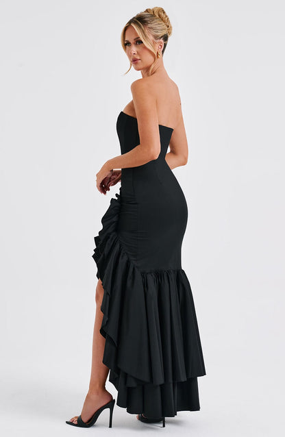Side profile of a woman wearing a strapless black Marlowe Ruffle Maxi Dress with a high-low ruffled hem, black heels.