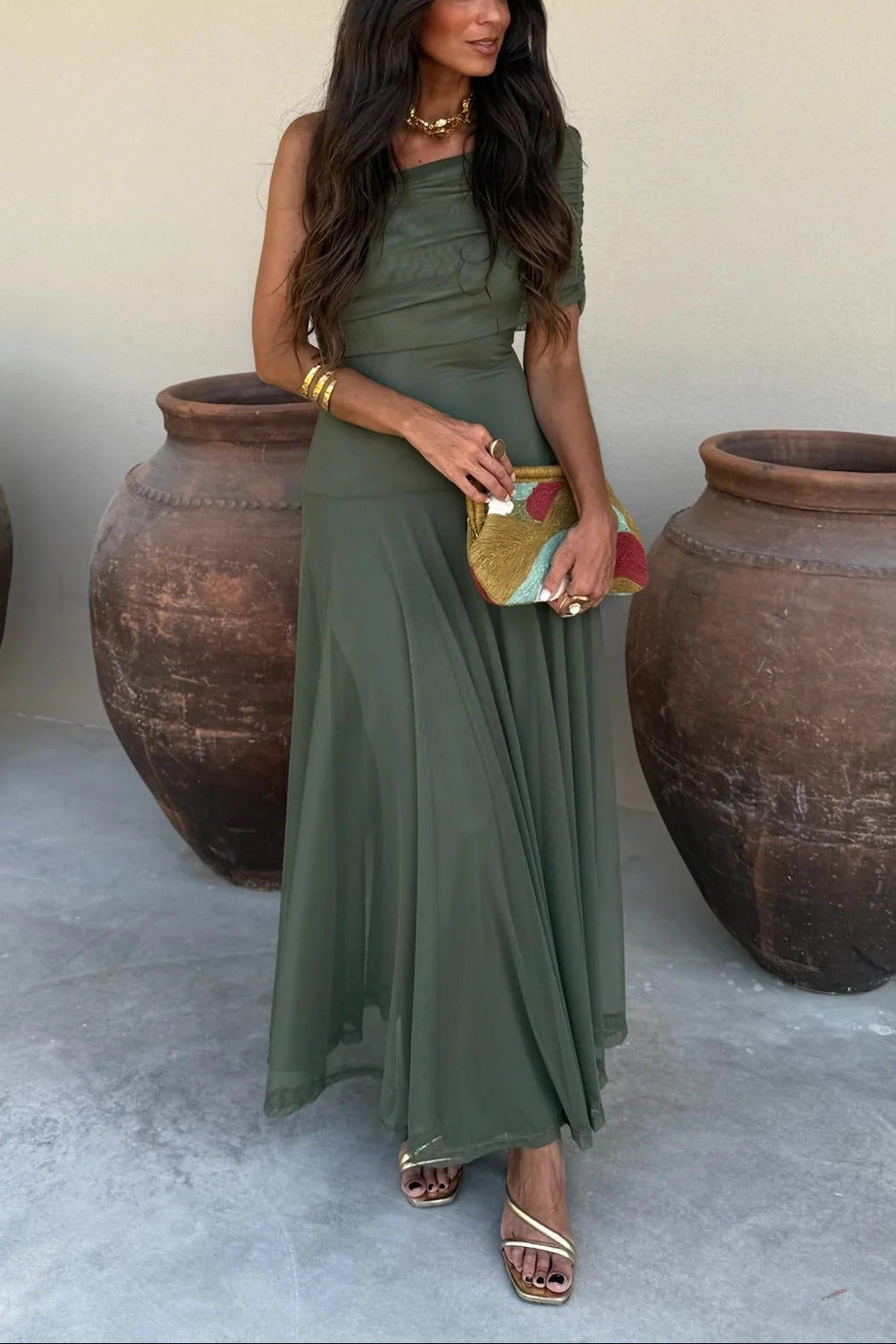 Milena draped maxi dress in olive green with a one-shoulder design, paired with gold heels and a colorful clutch.
