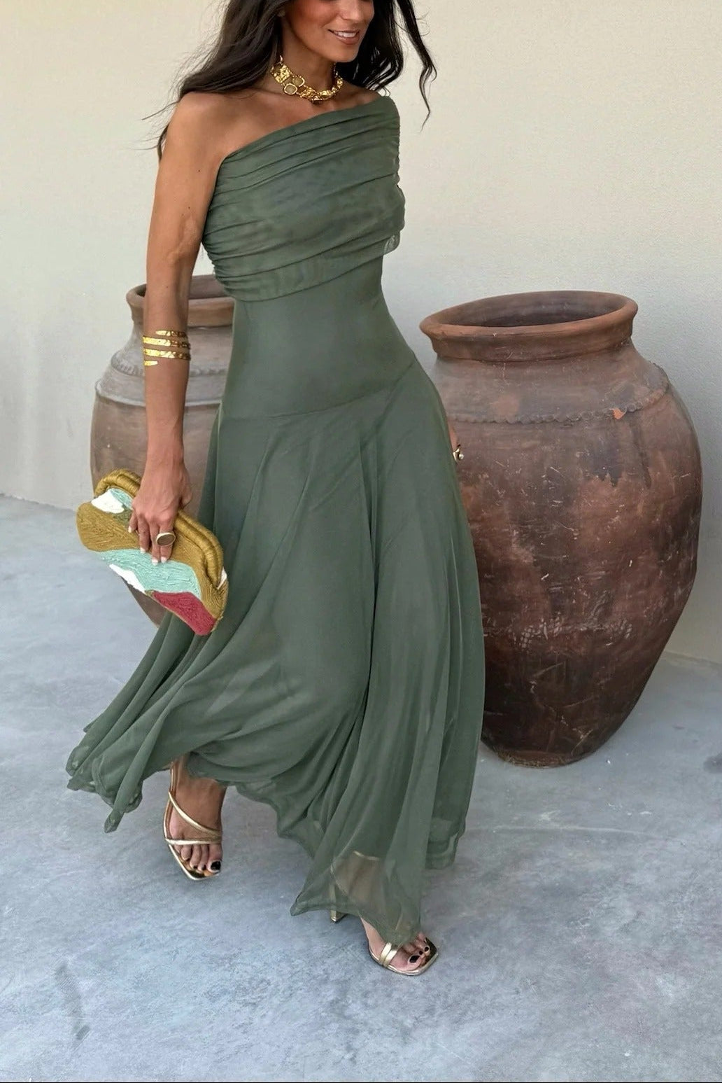 Full length view of a woman wearing a draped olive green maxi dress with one shoulder, gold heels, and a colorful clutch.