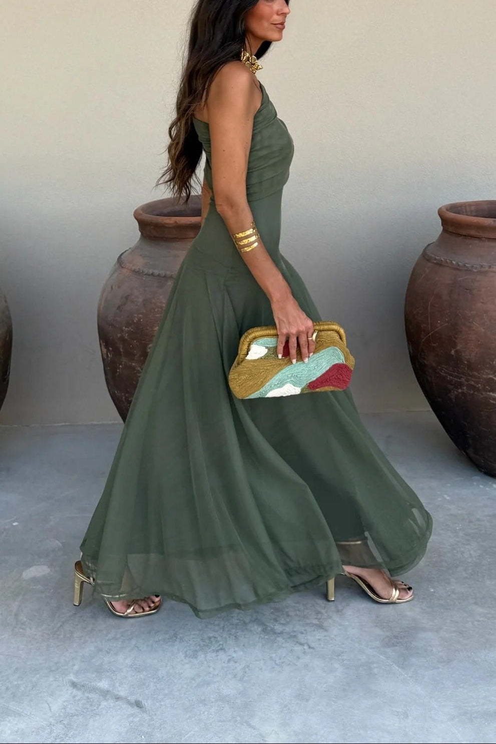 Side profile of a woman in an olive green draped maxi dress, showcasing the flowy skirt and a colorful clutch.