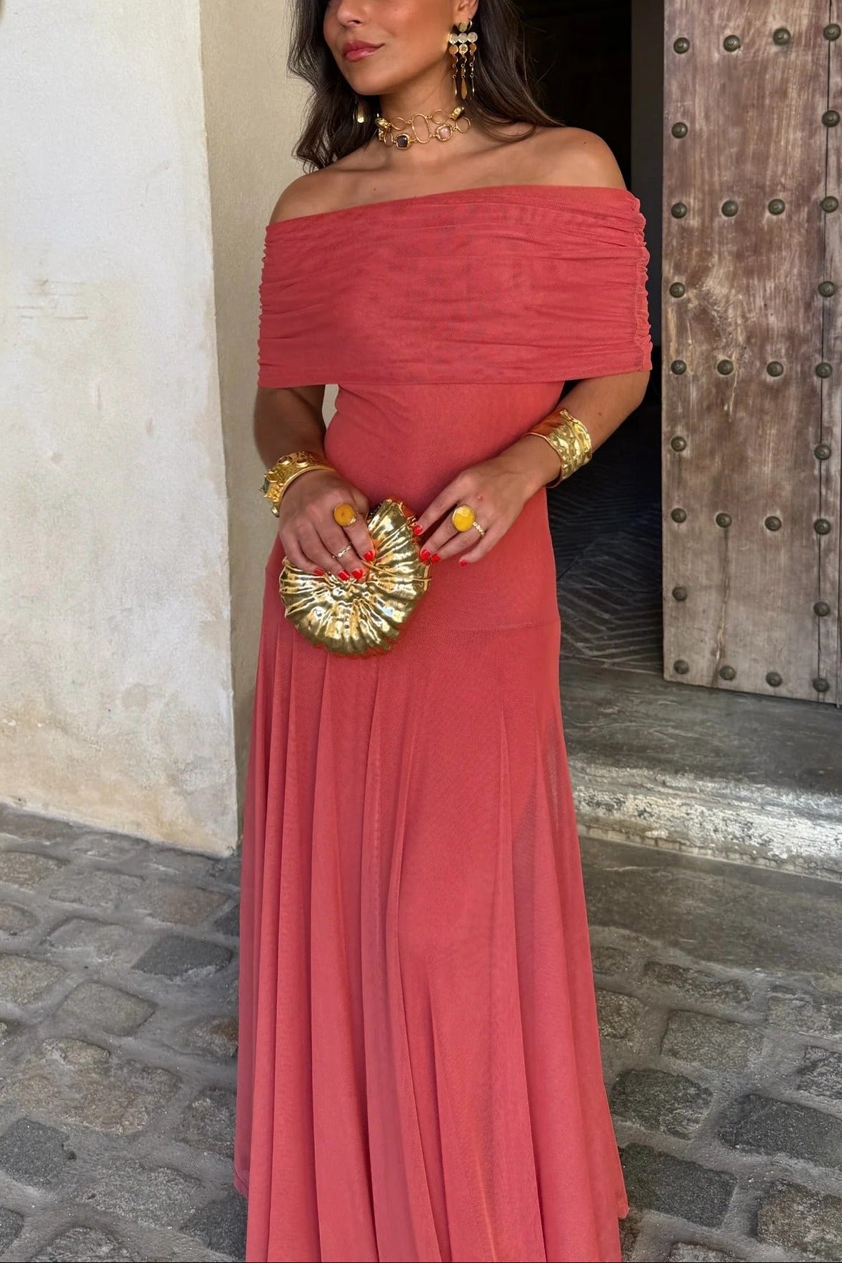 Milena draped maxi dress in a vibrant coral hue with an off-the-shoulder neckline and a golden clutch.