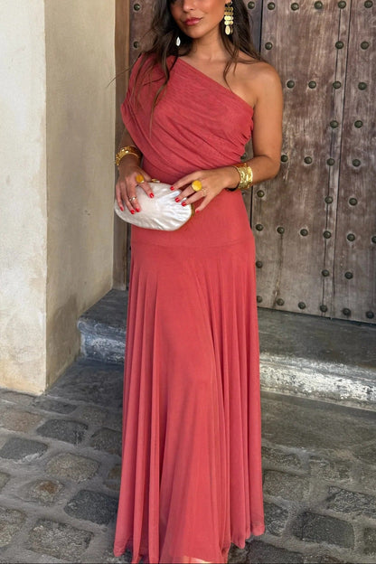 Front view of the coral Milena draped maxi dress, showcasing the one-shoulder style and a pearl clutch.