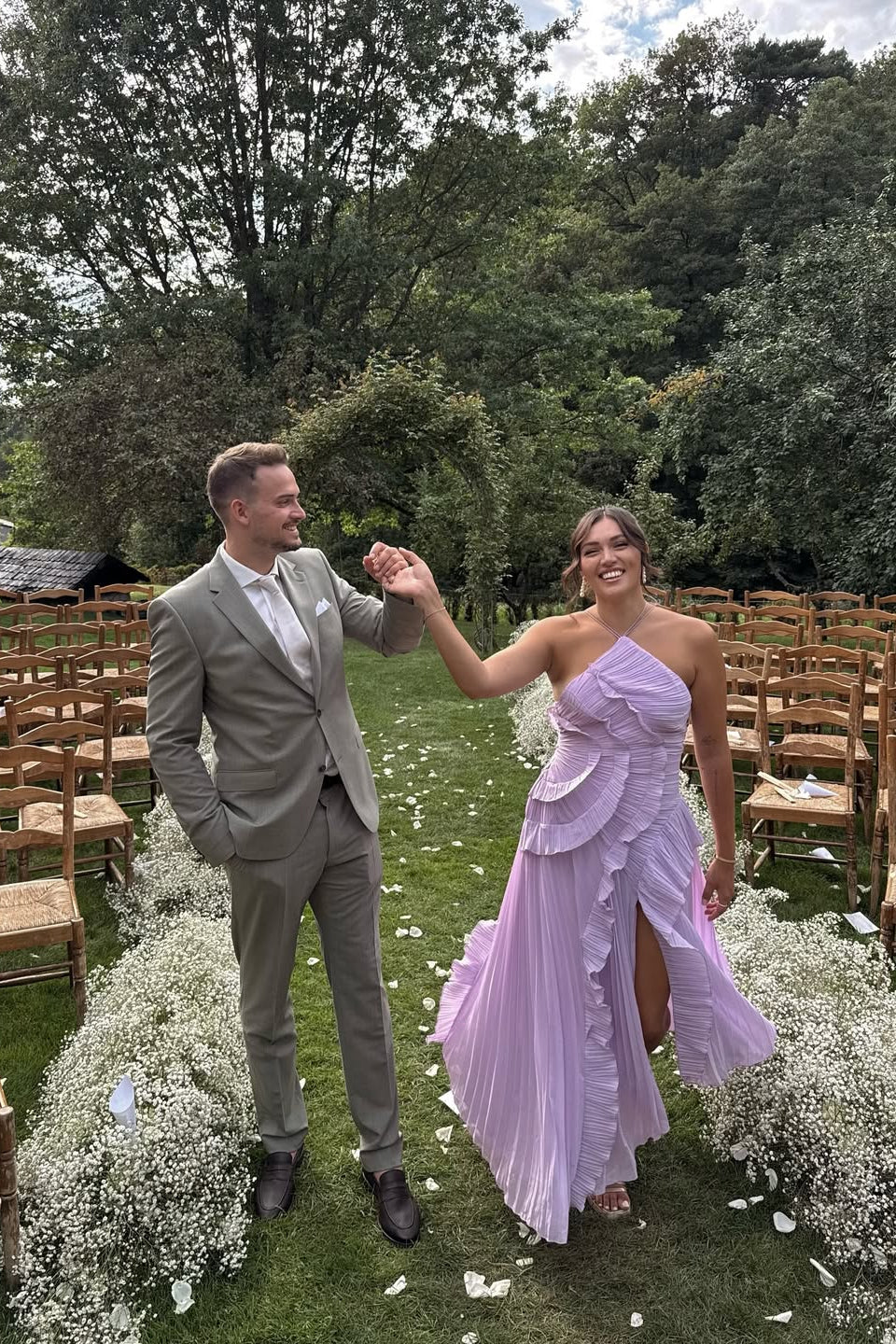 Woman in the purple Millie Ruffle Midi Dress with a high slit, holding hands with a man in a light grey suit, walking through an outdoor wedding aisle.