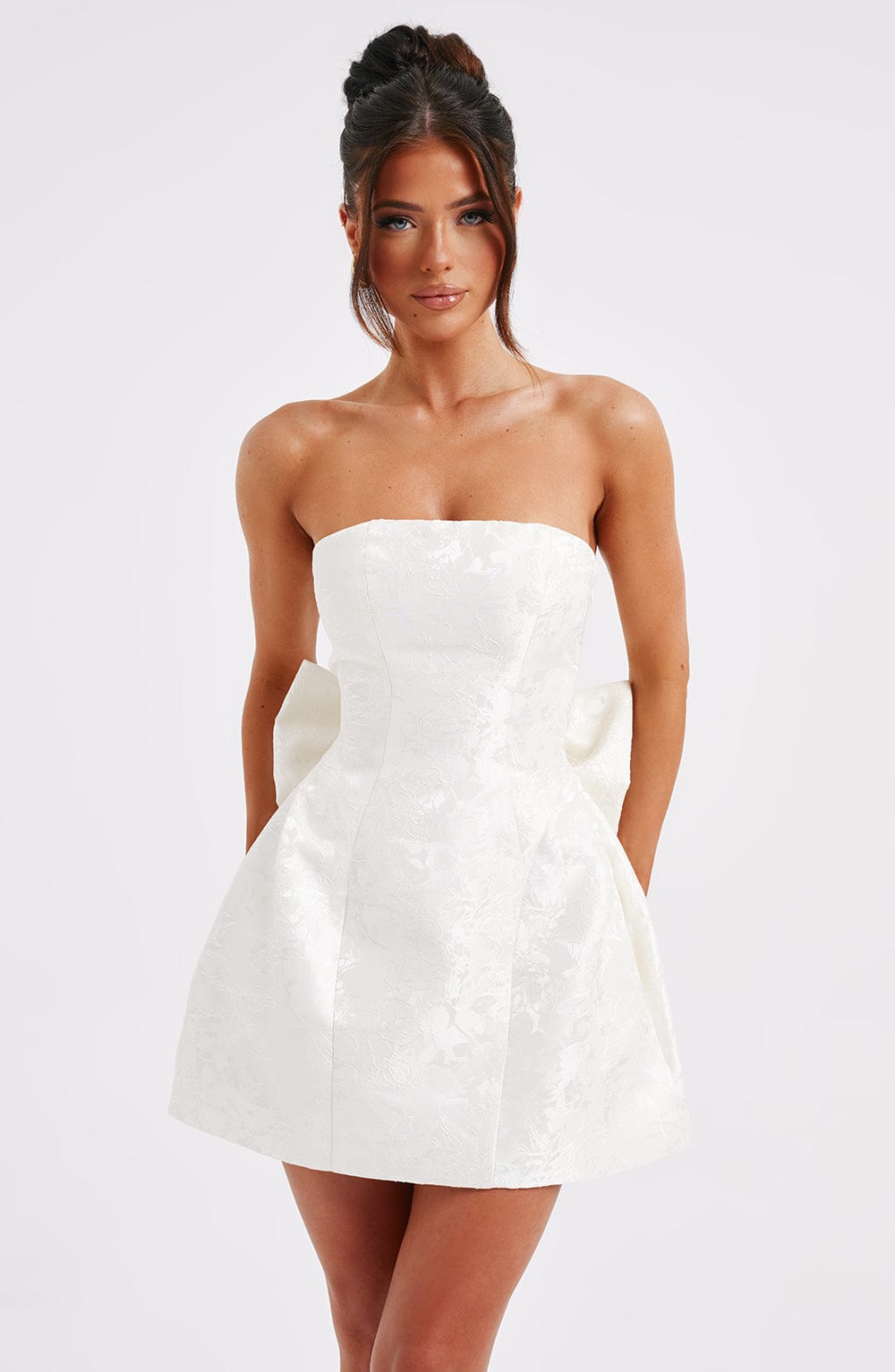 Front view of a white strapless mini dress with a subtle floral jacquard pattern and a large bow detail at the back.