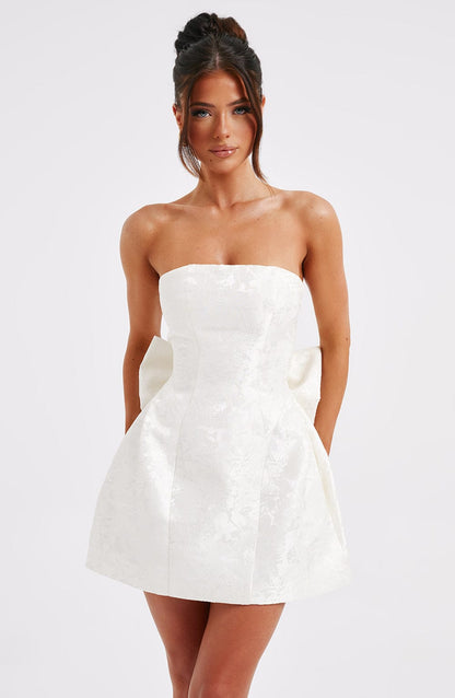 Front view of a white strapless mini dress with a subtle floral jacquard pattern and a large bow detail at the back.