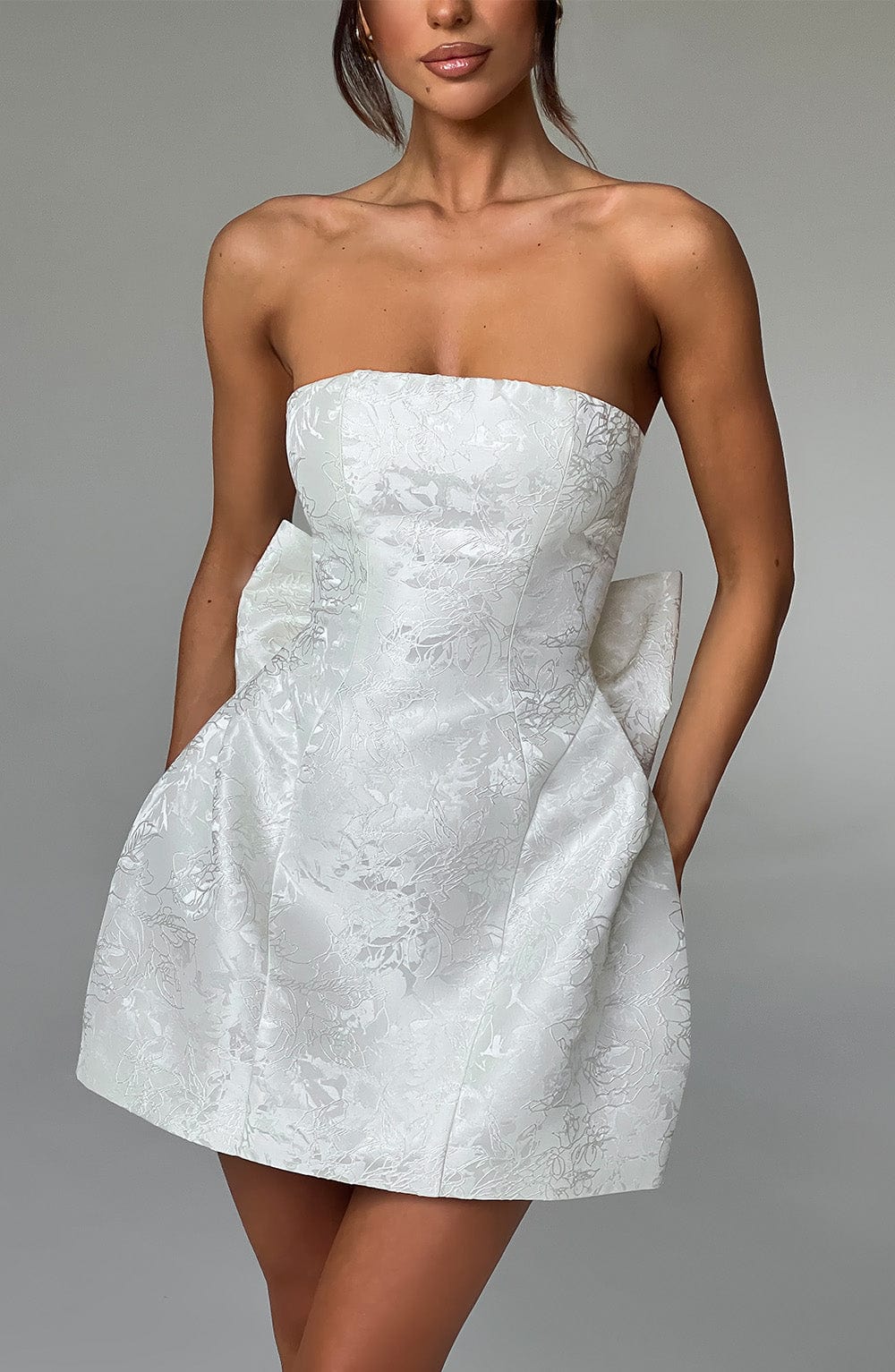 Angled front view of a white strapless mini dress with a floral jacquard pattern, featuring a large bow on the back.