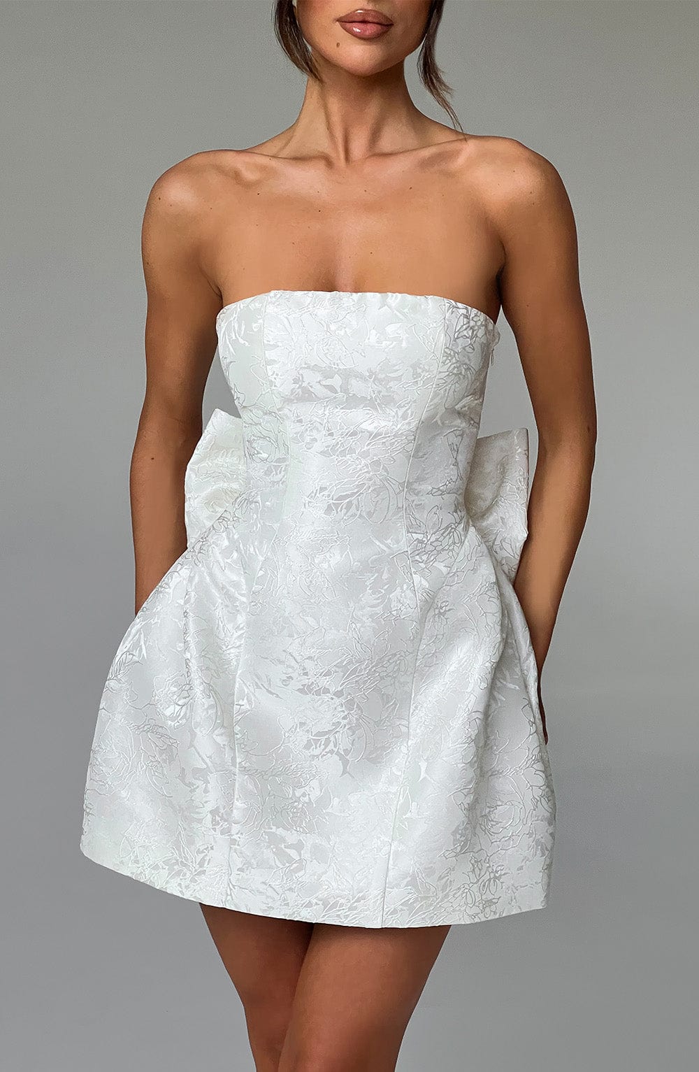Straight front view of a white strapless mini dress with a subtle floral jacquard pattern and a large bow on the back.