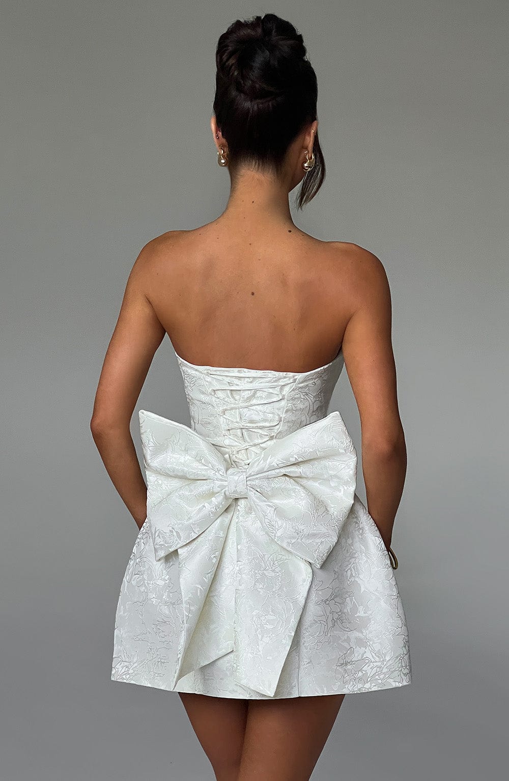 Back view of a white strapless mini dress with a lace-up corset back and a large elegant bow, perfect for wedding suits.