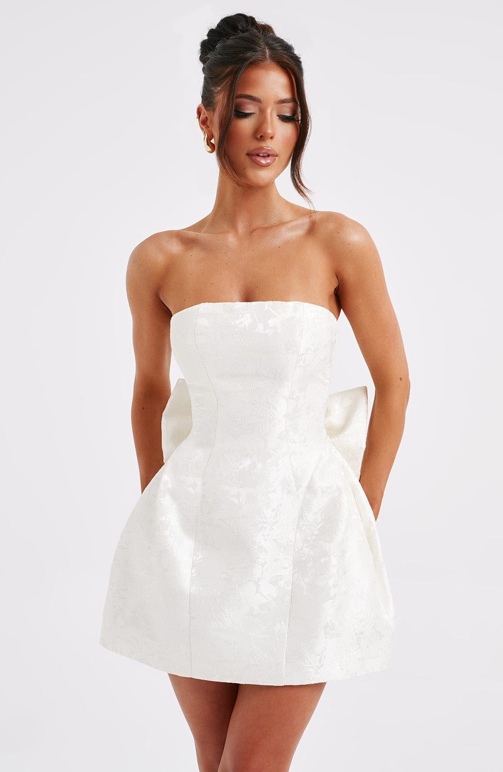 Close-up front view of a white strapless mini dress with a subtle floral jacquard fabric and a sculptural side bow, a sophisticated suit dress.