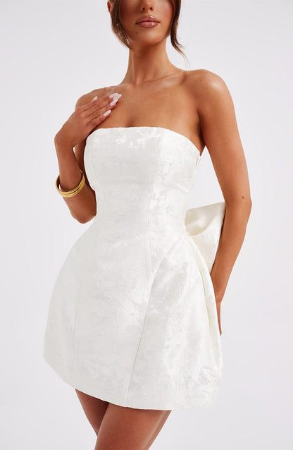 Side profile of a white strapless mini dress with a floral jacquard texture and a large bow, highlighting the tailored fit.