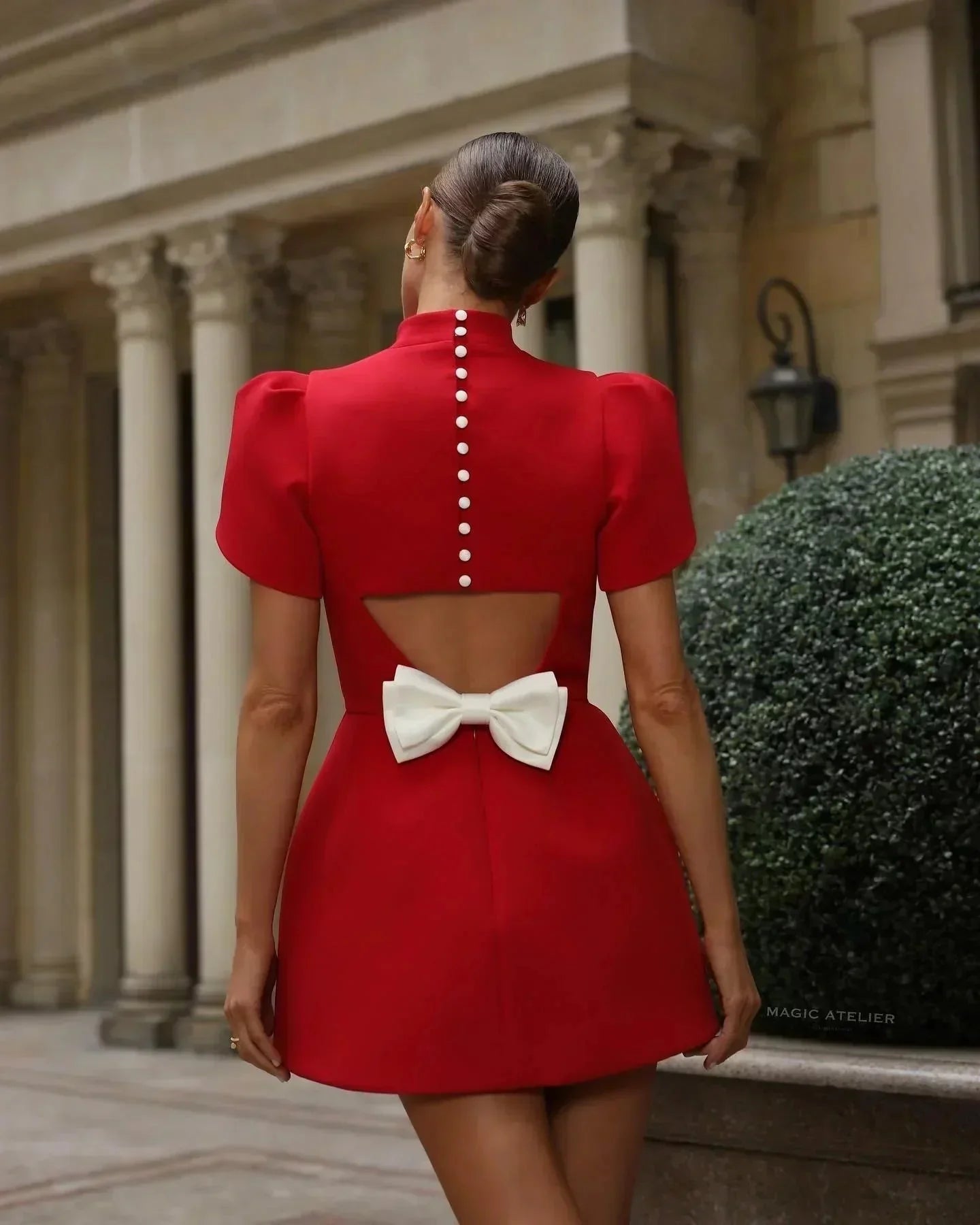 Full back view of the red Nora Bow Mini Dress with pearl buttons, open back, and a white bow, worn outdoors.