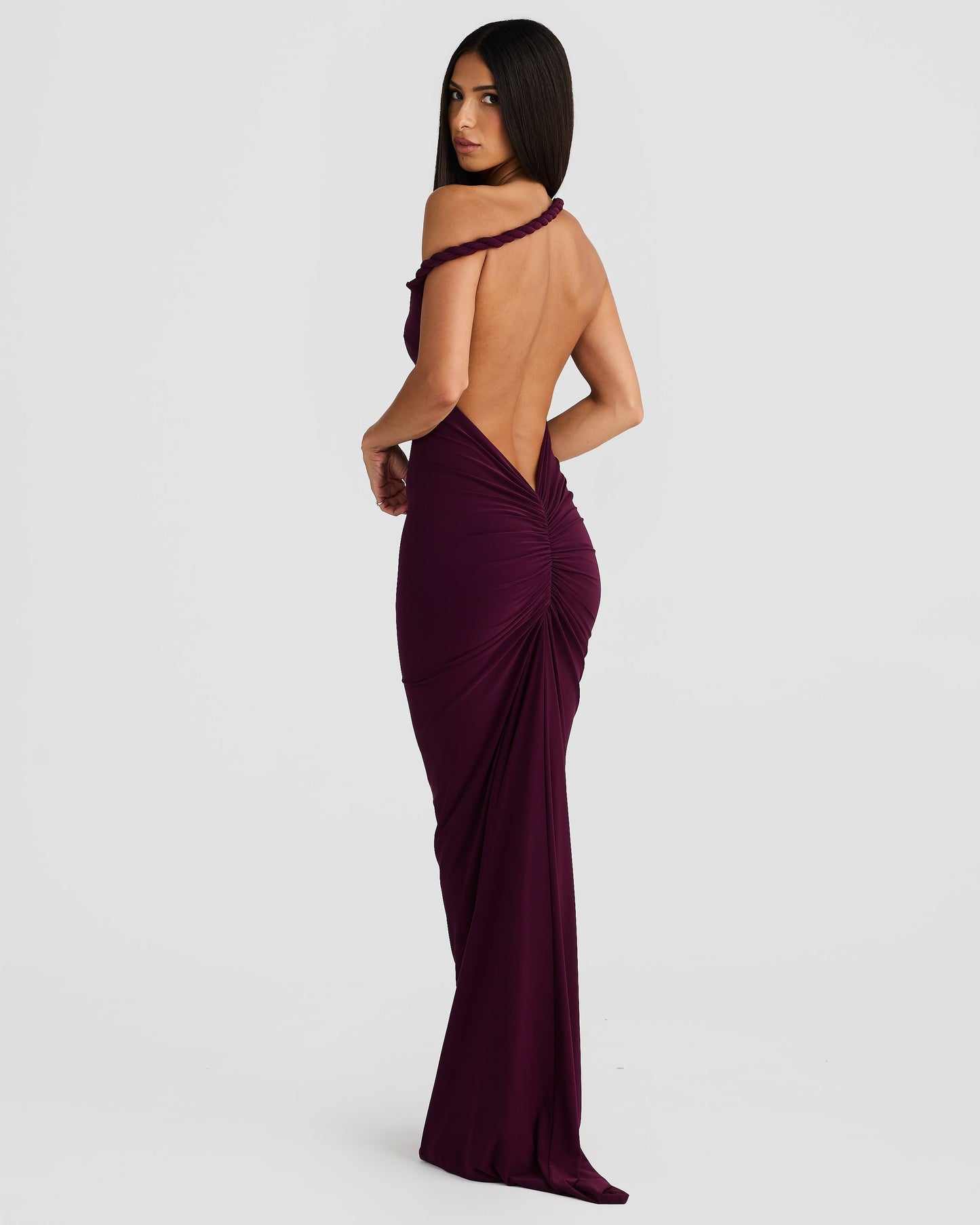 Side-back view of a woman in a plum purple backless maxi dress with a ruched back and one-shoulder detail.