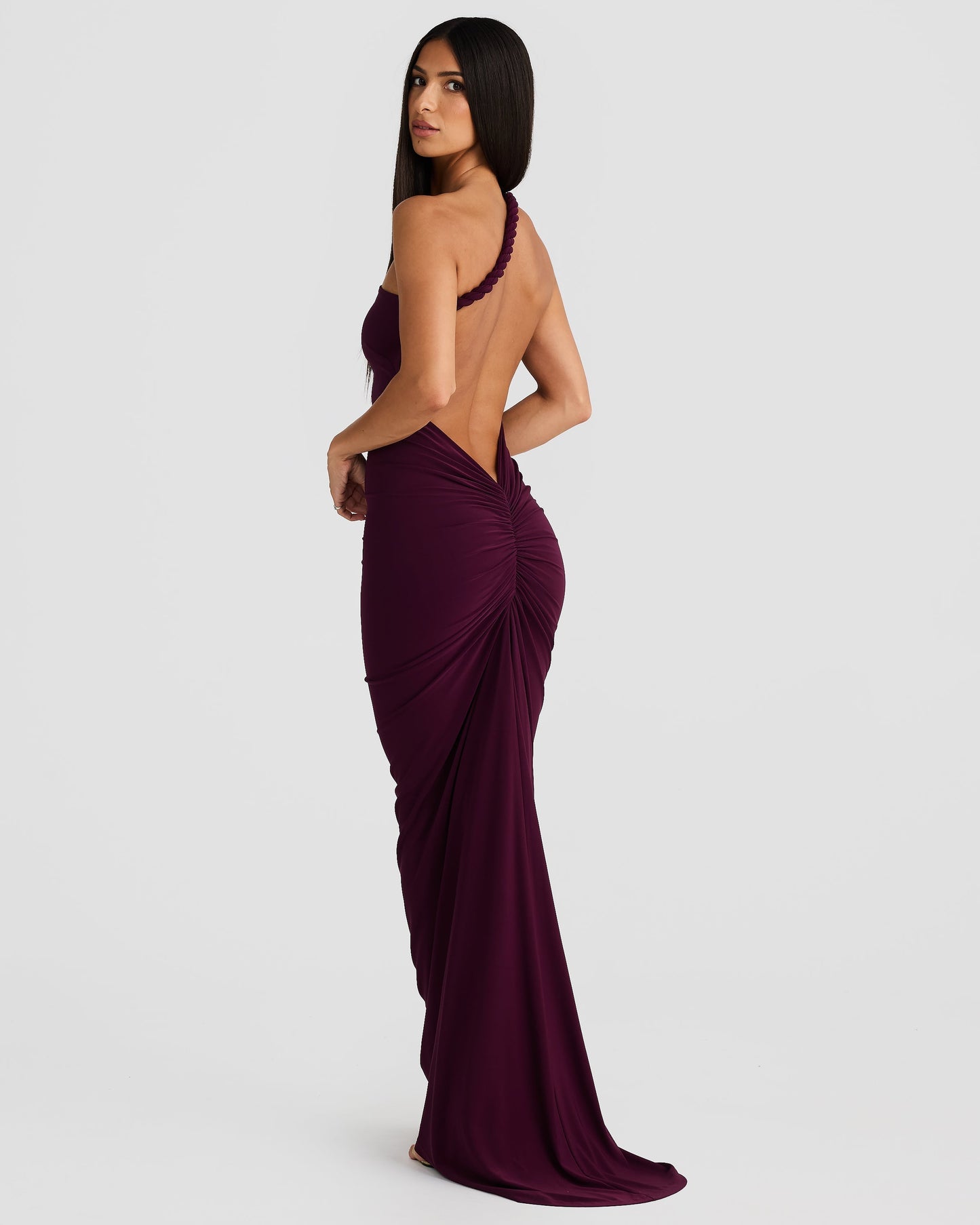 Side-back view of a woman in a plum purple Paulina backless maxi dress, showing the ruched back and braided strap.