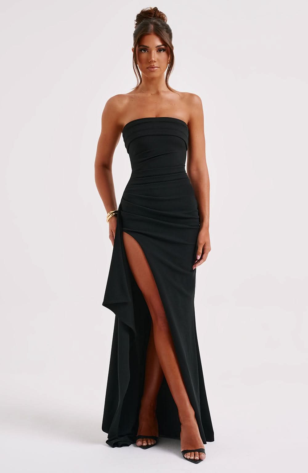 Front view of black strapless maxi dress featuring a ruched bodice, dramatic high slit, and elegant draped skirt.
