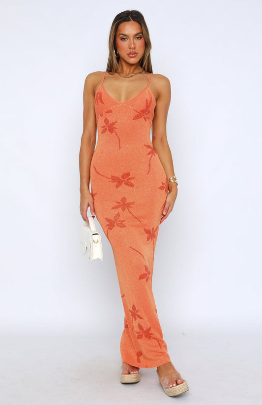 Full-length view of a woman wearing an orange Rebekah Floral Maxi Dress with spaghetti straps and subtle floral patterns, holding a white handbag and wearing espadrille slides.