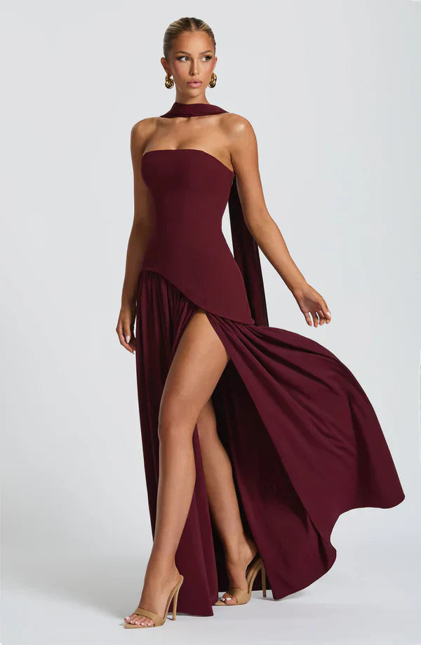 Burgundy strapless maxi dress with a high leg slit, asymmetrical peplum, and a flowing scarf detail, in motion.