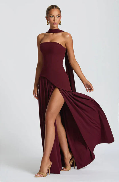 Burgundy strapless maxi dress with a high leg slit, asymmetrical peplum, and a flowing scarf detail, in motion.
