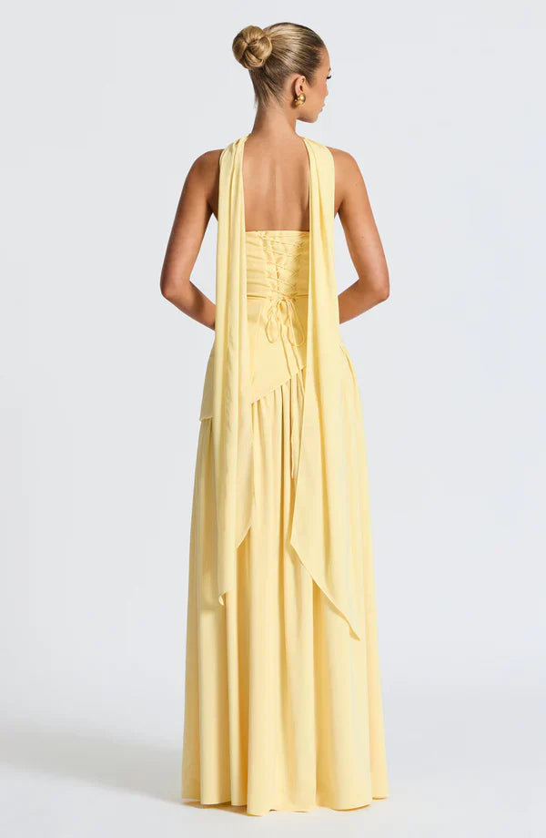 Back view of the light yellow Rhea Asymmetrical Maxi Dress, showing the intricate corset lacing and long draped fabric.