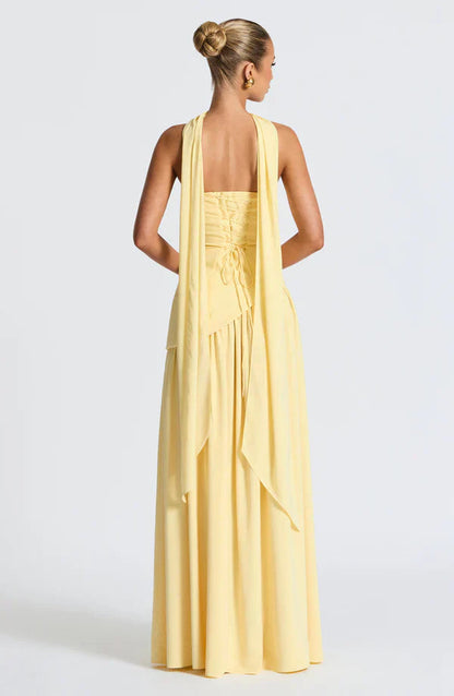 Back view of the light yellow Rhea Asymmetrical Maxi Dress, showing the intricate corset lacing and long draped fabric.