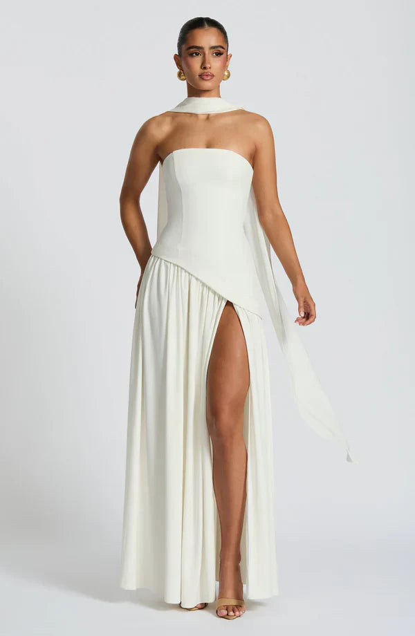 Elegant white strapless maxi dress with a high leg slit, asymmetrical peplum, and a long flowing scarf detail.