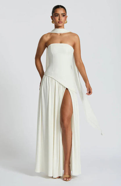 Elegant white strapless maxi dress with a high leg slit, asymmetrical peplum, and a long flowing scarf detail.