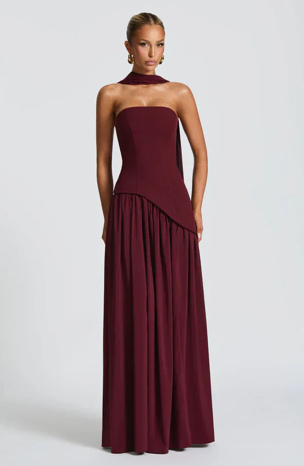 Deep burgundy strapless maxi dress with asymmetrical peplum waist and flowing skirt, featuring a matching choker detail.