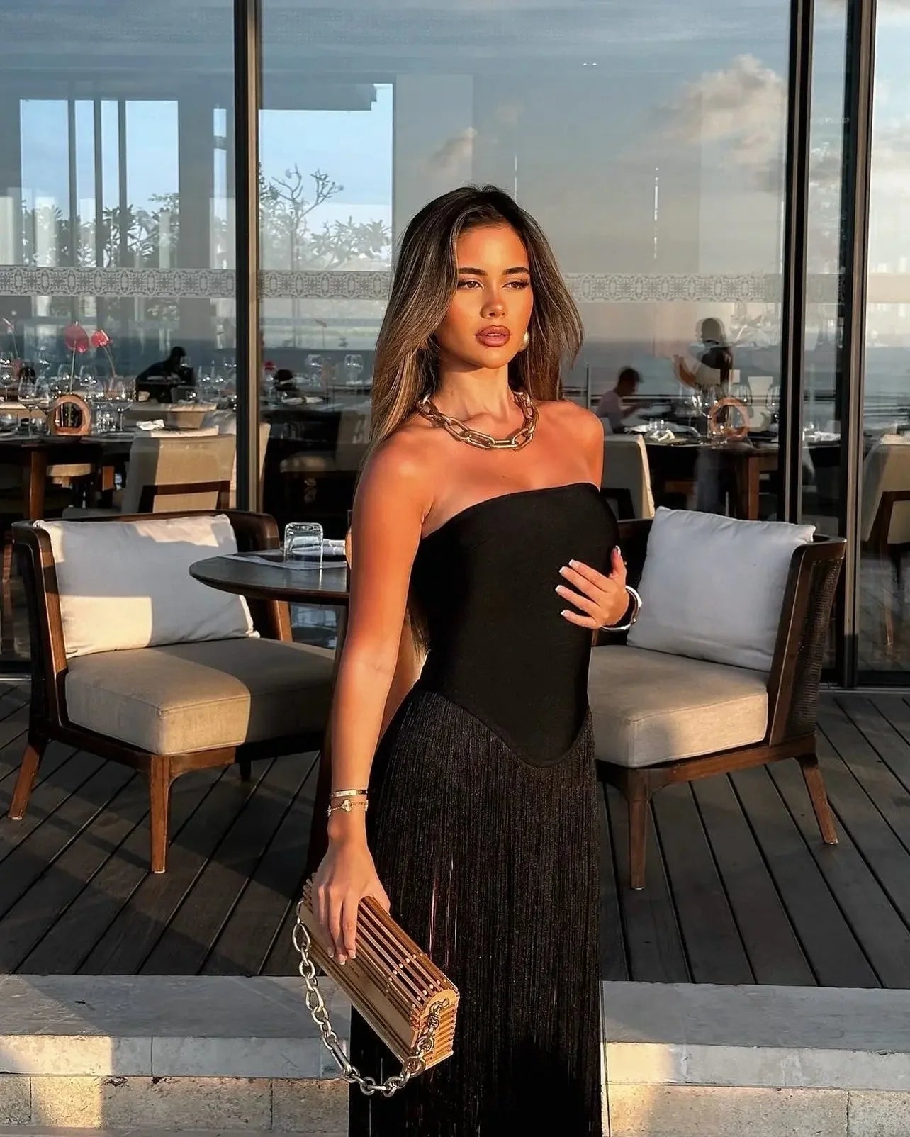 Woman in a black strapless Adaline Fringe Maxi Dress with a structured bodice and long fringe skirt, holding a wooden clutch, standing on a patio.