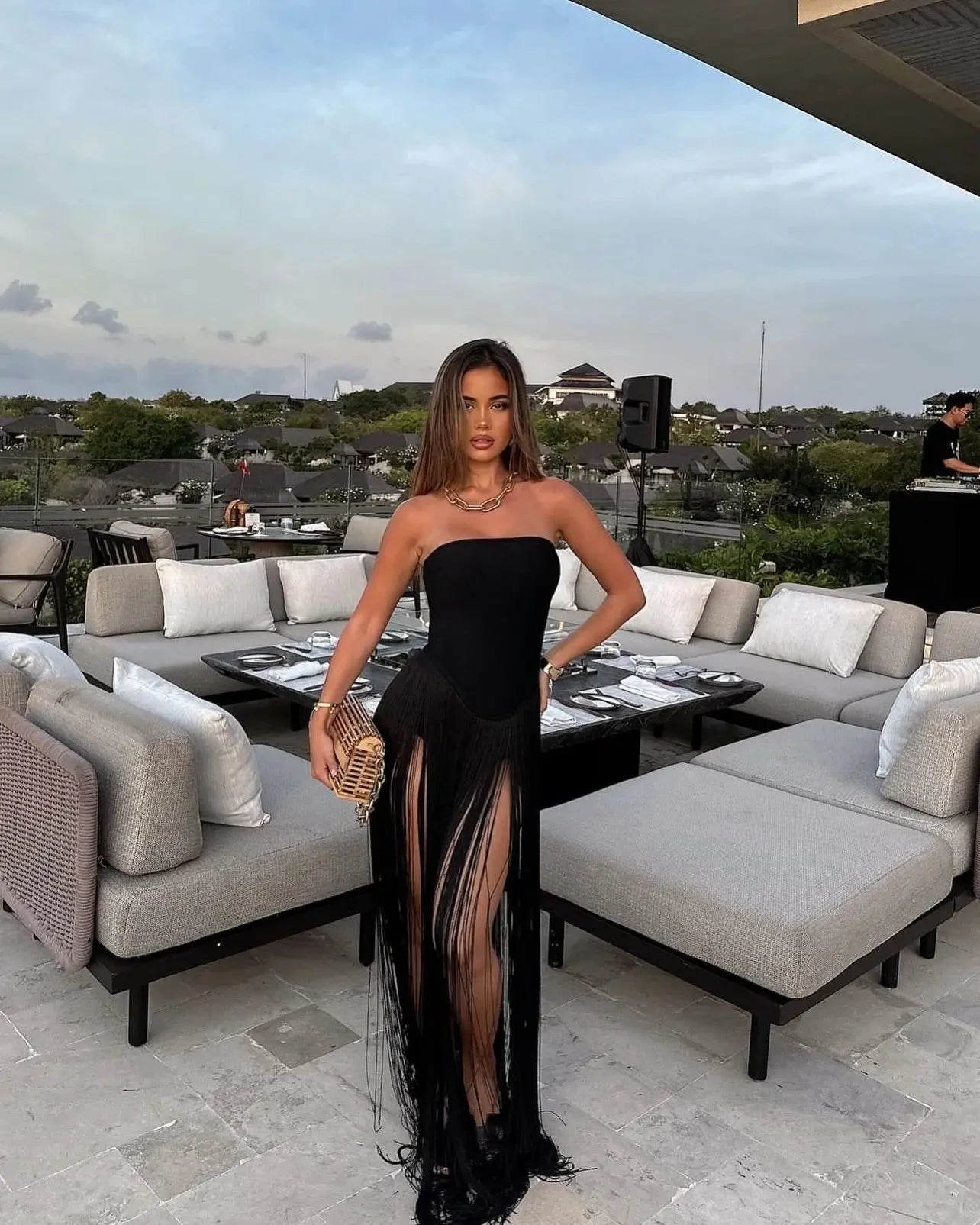 Full-length view of a woman wearing a black strapless Adaline Fringe Maxi Dress with a high leg slit and flowing fringe detail, posing outdoors.