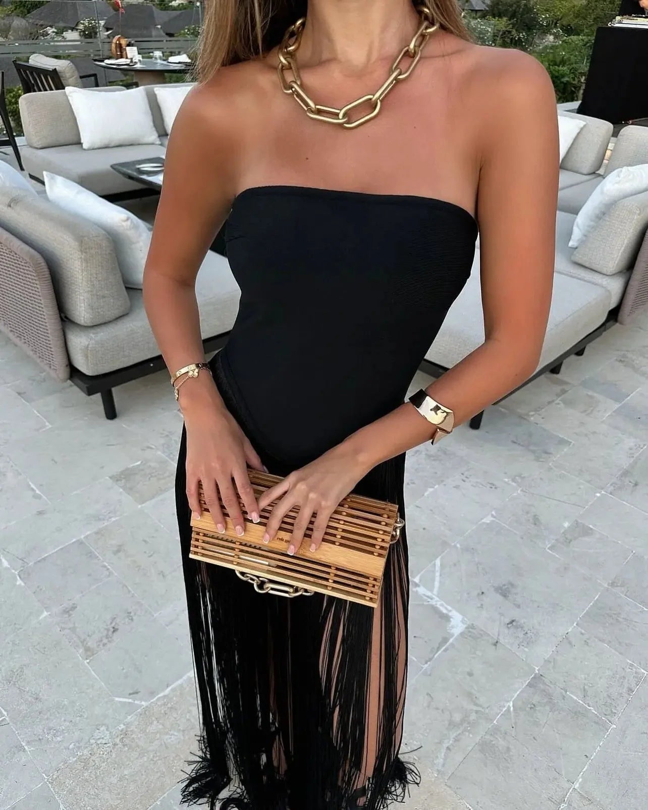 Close-up of the black strapless Adaline Fringe Maxi Dress bodice and flowing fringe skirt, paired with a chunky gold necklace and wooden clutch.
